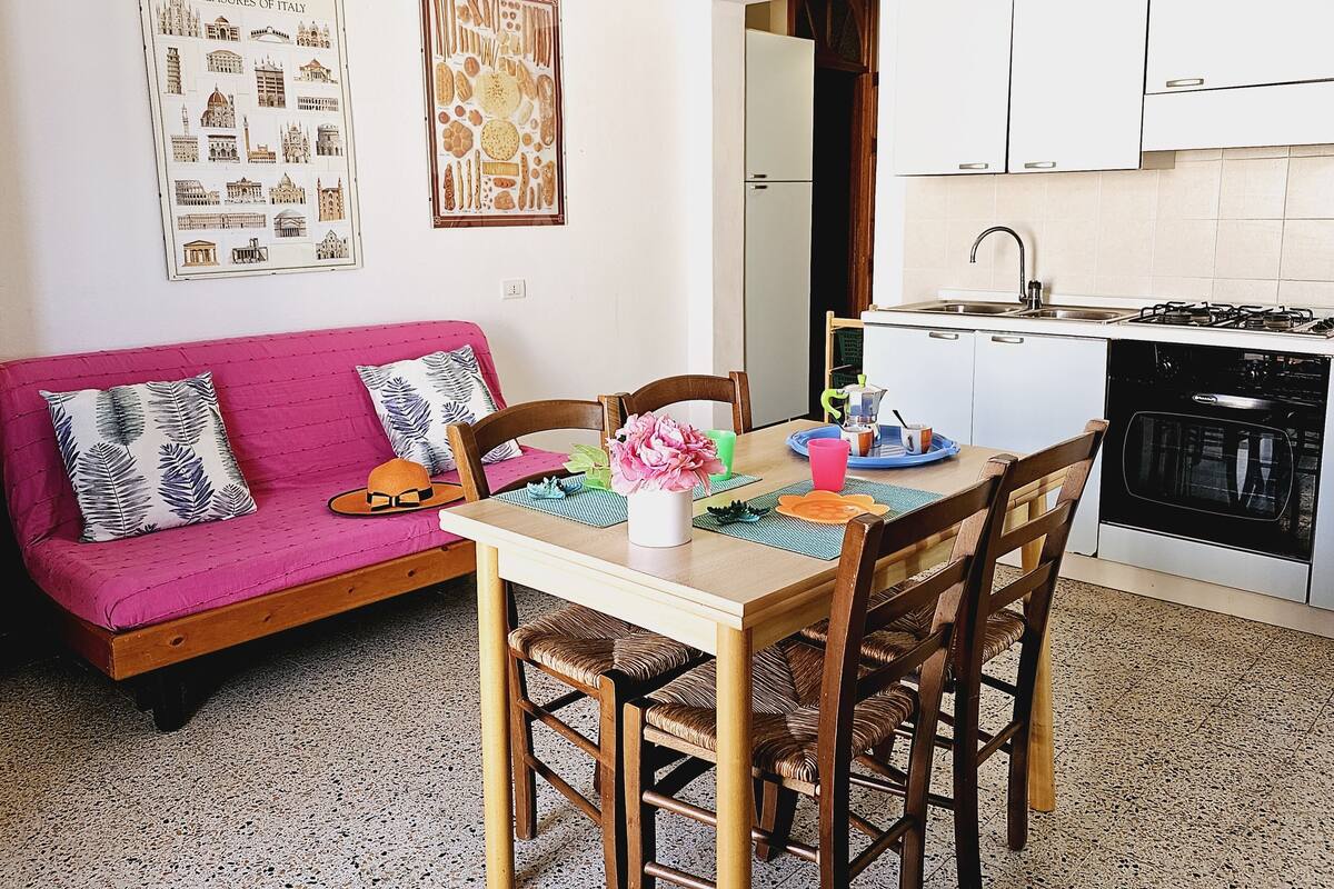 Une cuisine chaleureuse avec une table à manger, des chaises et un canapé rose. On y trouve des tableaux encadrés au mur, un évier et une cuisinière.