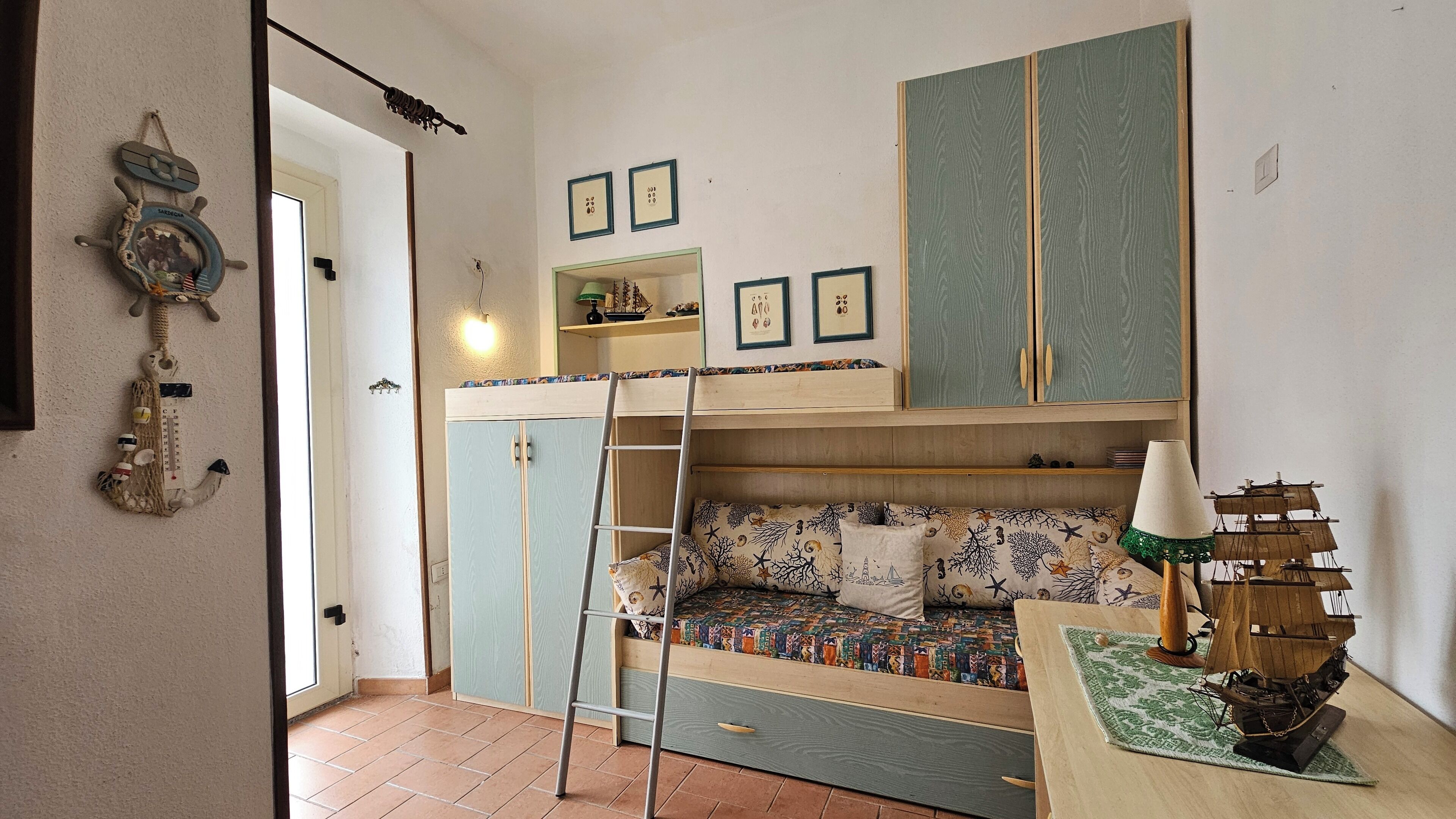 Une petite pièce comprenant un lit superposé, une échelle, une petite table sur laquelle est posé un modèle de bateau, et des décorations sur le thème de la marine.
