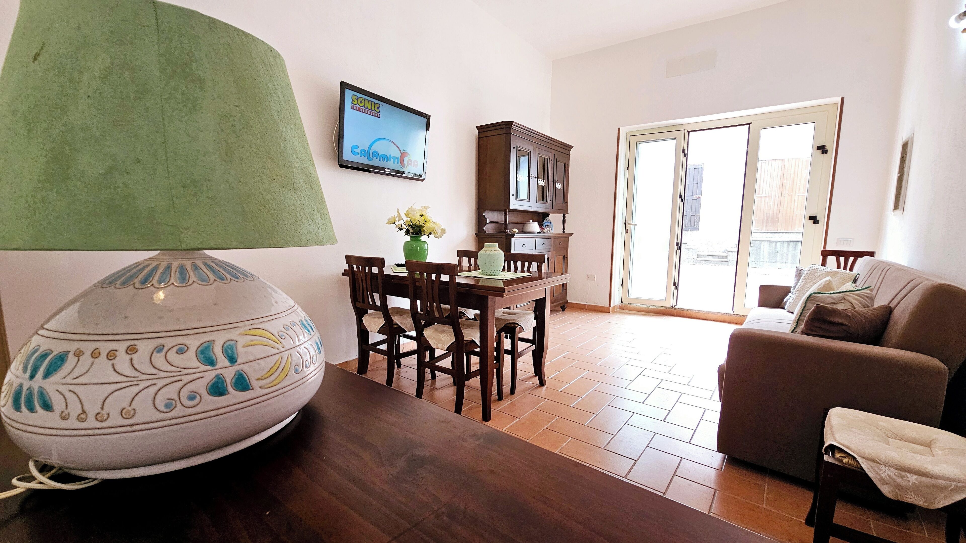 A living room with a wooden table, chairs, a sofa, a lamp, and a TV displaying 'Sonic the Hedgehog'.