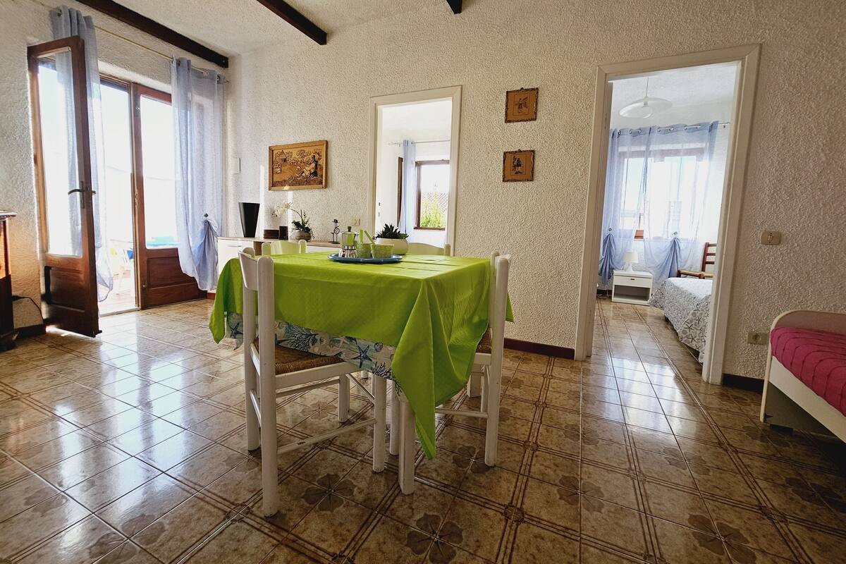 Un espace repas avec une table verte, des chaises blanches et un sol carrelé. Deux portes donnent accès à d’autres pièces.
