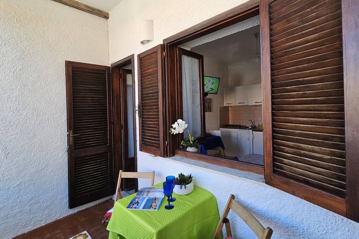 Un petit coin repas avec une table dressée pour deux personnes, une chaise, une fenêtre avec des volets en bois et une vue sur la cuisine.