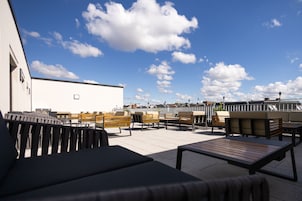 An outdoor seating area with wooden and metal furniture, a white building in the background, and a clear blue sky with clouds.