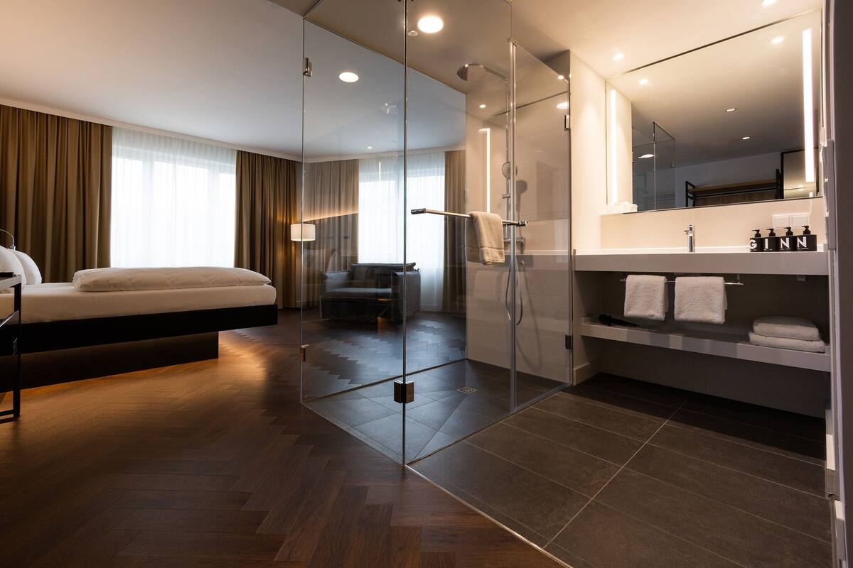 A modern hotel room with a bed, a glass-enclosed shower, a vanity with a mirror, and a large window with curtains.