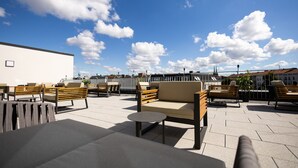 Un espacio exterior con varios sofás y sillones, una pequeña mesa redonda y vistas a la ciudad al fondo.