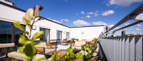 A rooftop terrace with outdoor seating, a view of the city skyline, and a clear blue sky.