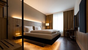 Una habitación de hotel moderna con una cama suspendida, suelo de madera, una mesita de noche, un televisor y un amplio ventanal con cortinas.