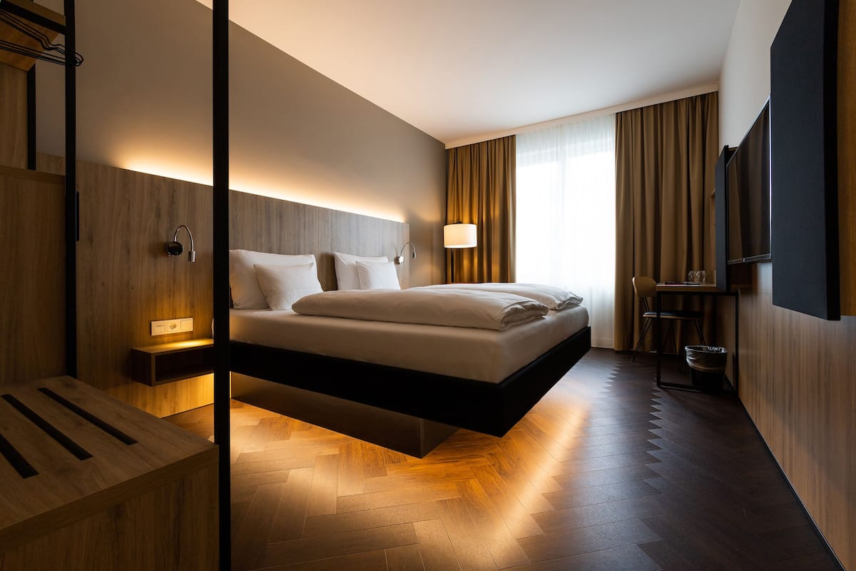 A modern hotel room with a suspended bed, wooden flooring, a bedside table, a TV, and a large window with curtains.
