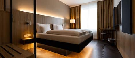 A modern hotel room with a suspended bed, wooden flooring, a bedside table, a TV, and a large window with curtains.