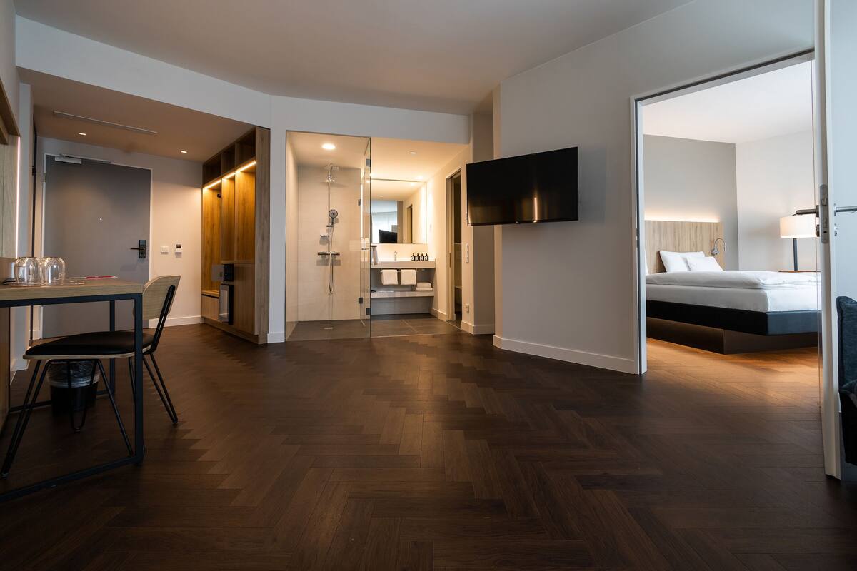 A modern hotel room with a wooden floor, a bed, a TV mounted on the wall, and a bathroom visible through an open door.