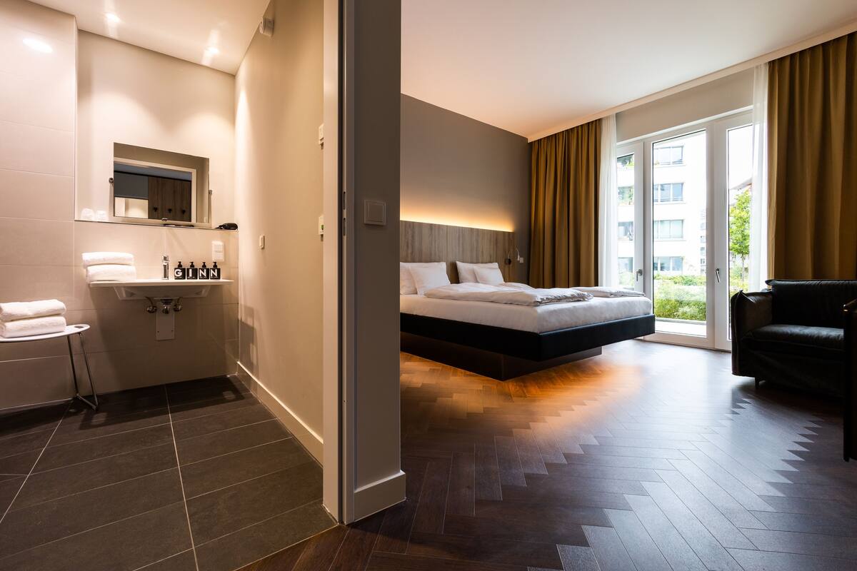 A modern hotel room with a large bed, a separate sitting area, a bathroom with a sink and mirror, and a view of the outdoors through a sliding door.