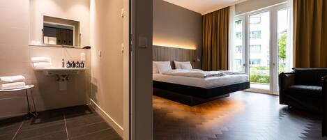 A modern hotel room with a large bed, a separate sitting area, a bathroom with a sink and mirror, and a view of the outdoors through a sliding door.