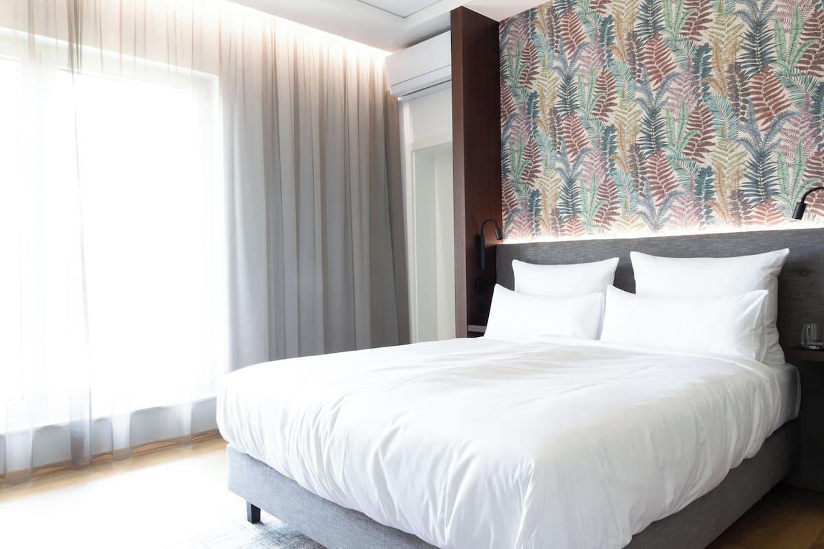 A modern hotel room with a large bed, a headboard with a patterned wallpaper, and a large window with sheer curtains.
