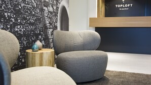 Un hall d’hôtel moderne avec des sièges gris à revêtement en tissu, une table d’appoint en bois agrémentée de vases décoratifs et un mur orné d’un motif abstrait en noir et blanc.