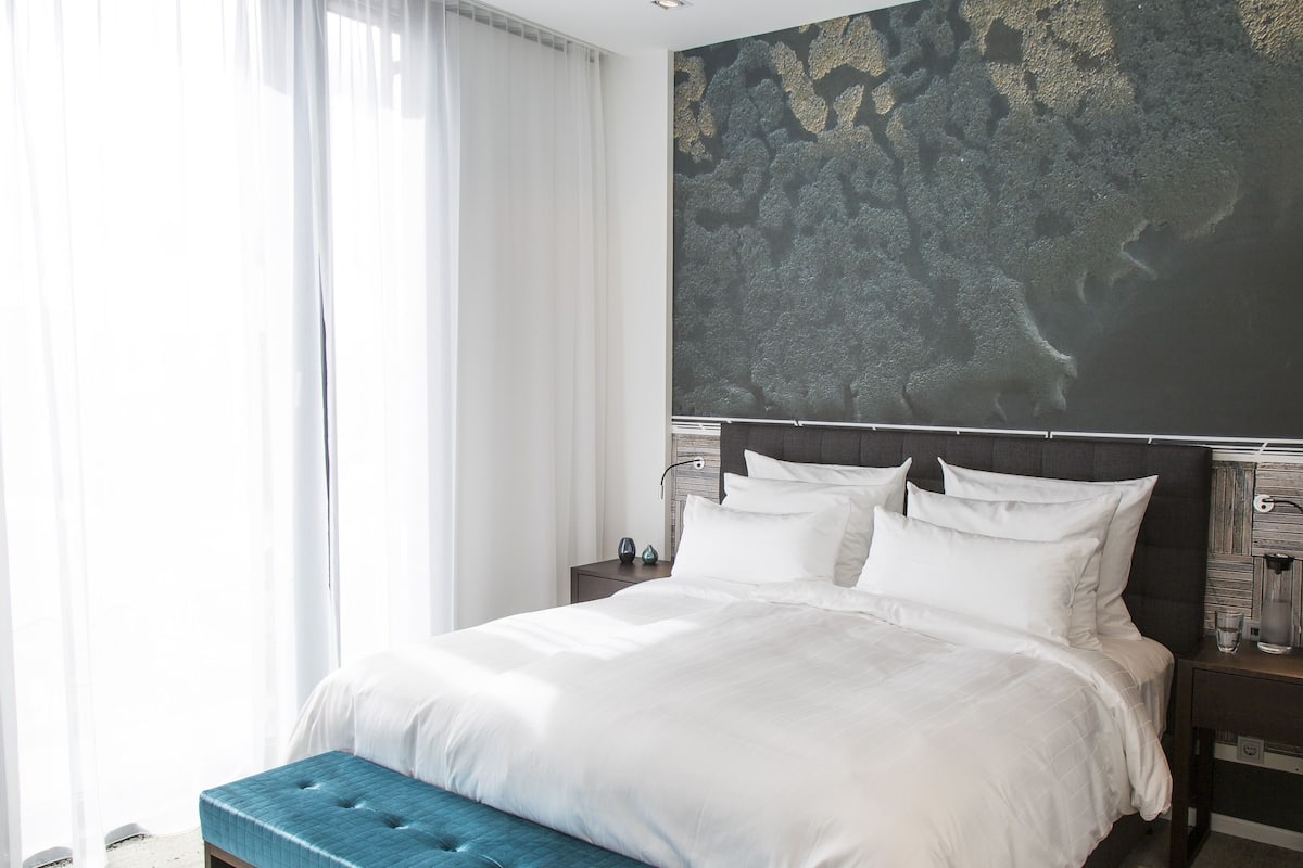 A modern hotel room with a large bed, a blue bench, a nightstand with a lamp, and a wall with a textured design.