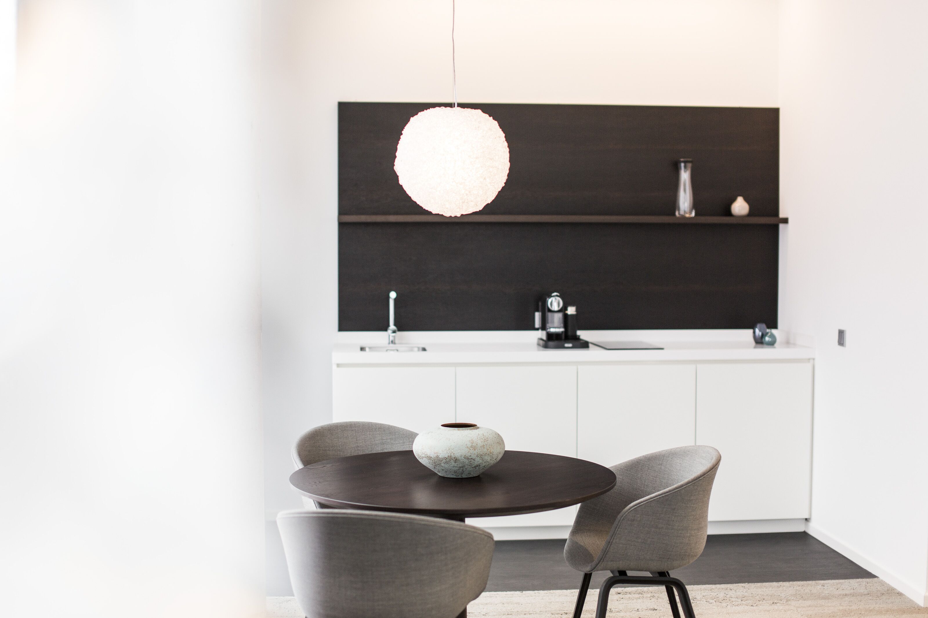 A modern kitchen with a dark wooden upper shelf, a white countertop, and a round dining table with two chairs.