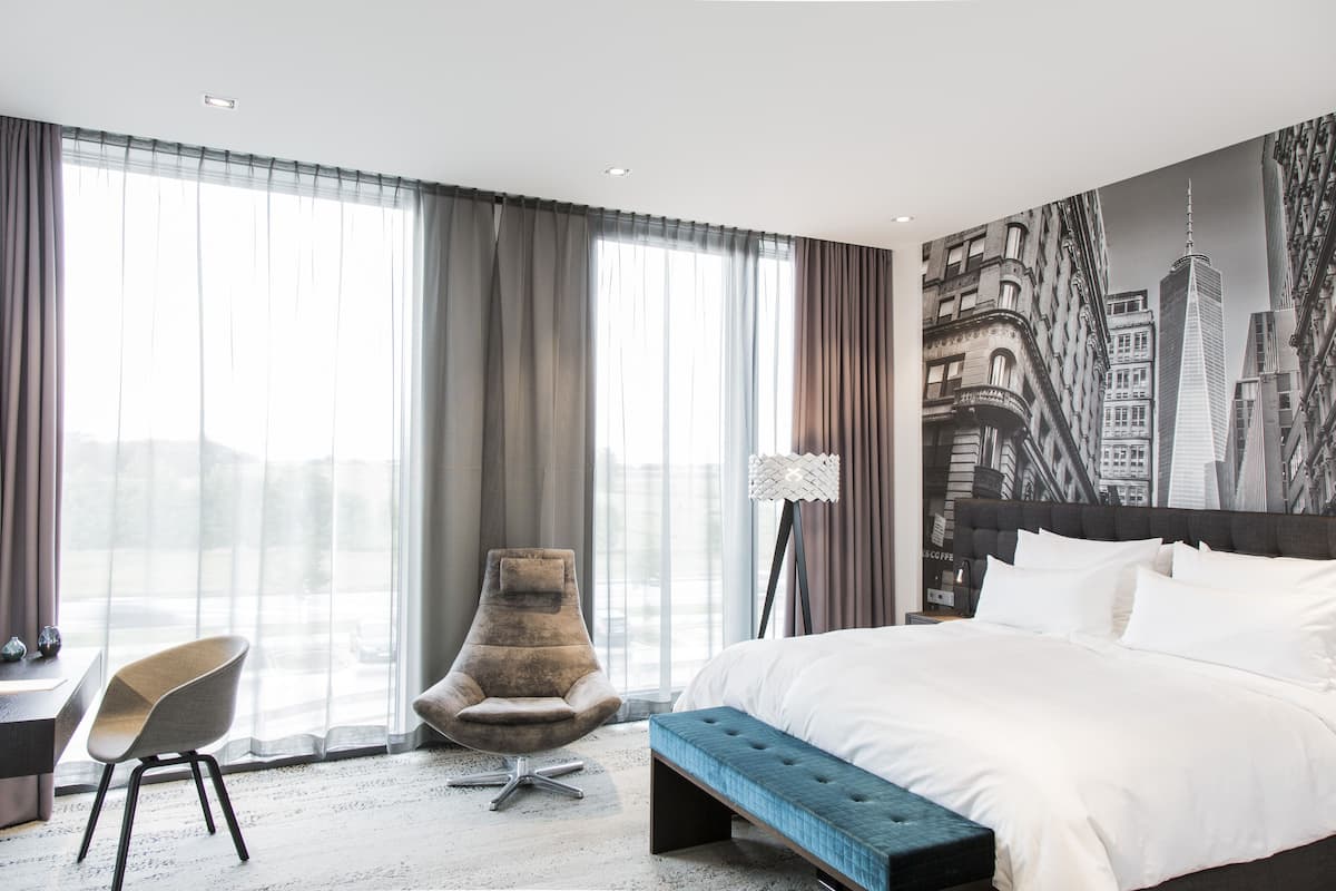 A modern hotel room with a large bed, a chair, a bench, and a cityscape mural on the wall.