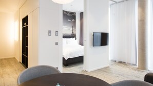 Une chambre d’hôtel moderne équipée d’un lit, d’un téléviseur à écran plat, d’un coin repas avec une table et des chaises, et d’une grande fenêtre avec des rideaux.
