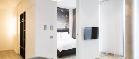 Una habitación de hotel moderna con cama, televisor de pantalla plana, zona de comedor con mesa y sillas, y un amplio ventanal con cortinas.