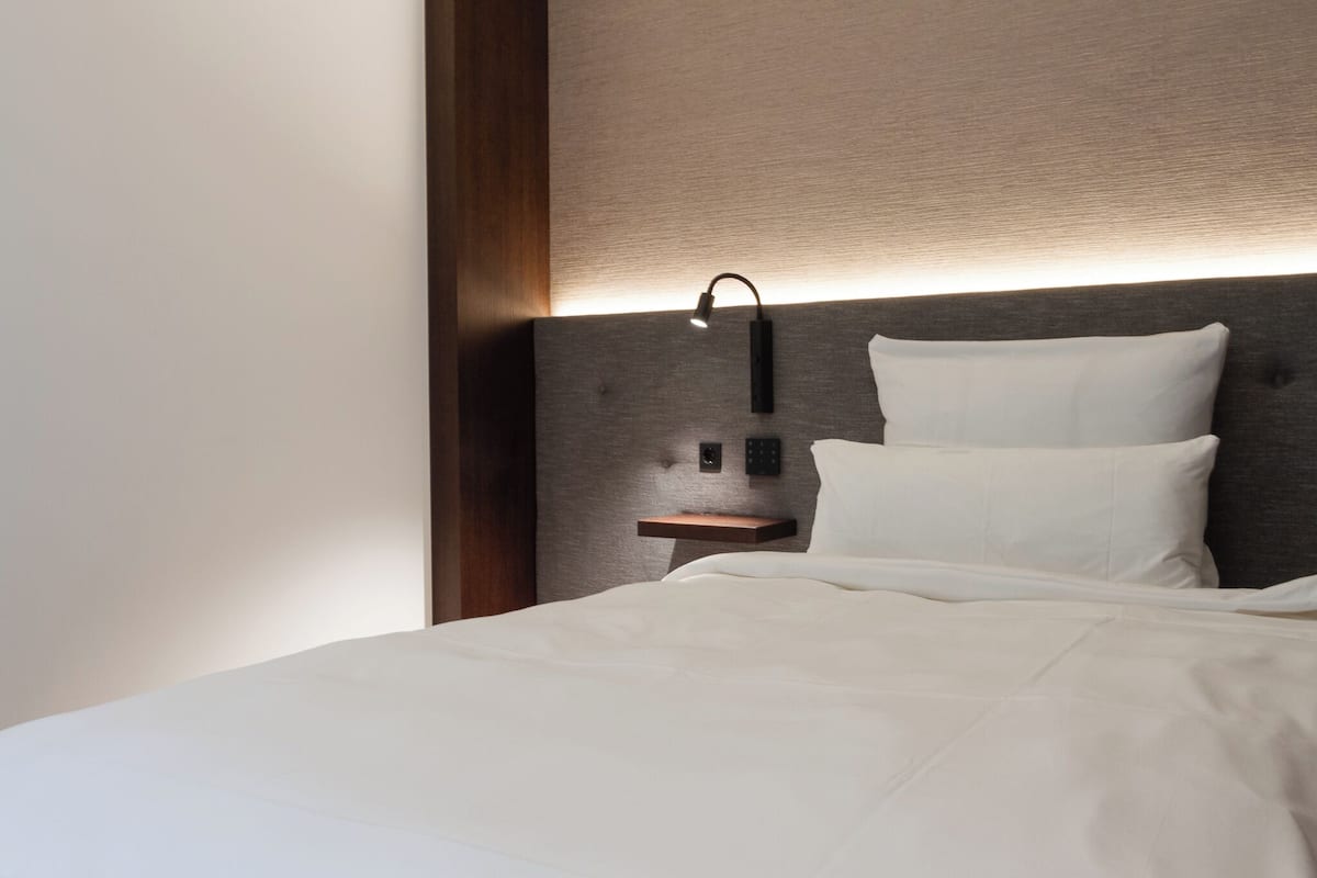 A neatly made bed with white linens, a wall-mounted nightstand with a lamp, and a textured wall behind the bed.