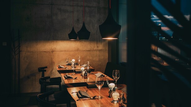 Un restaurant à la lumière tamisée, avec une longue table en bois dressée pour le dîner, des lampes suspendues et une vue sur le paysage urbain par une fenêtre.