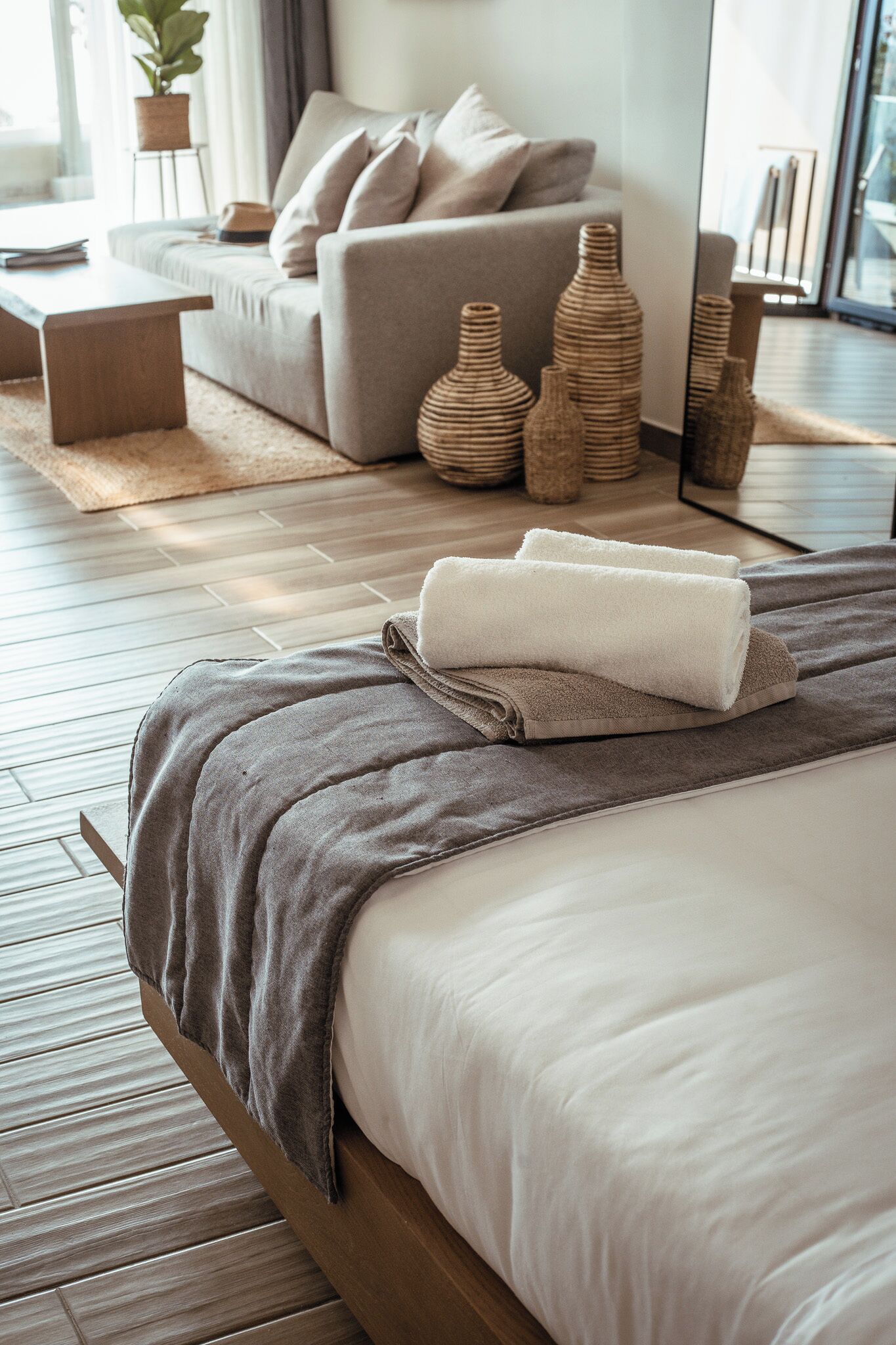 A neatly made bed with a grey blanket and white towels, a wooden headboard, a grey sofa with cushions, and a small table with a plant.