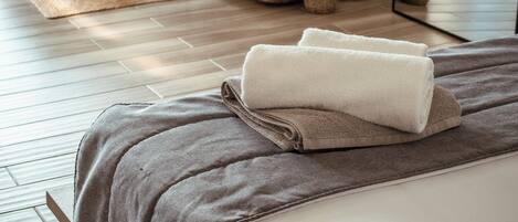 A neatly made bed with a grey blanket and white towels, a wooden headboard, a grey sofa with cushions, and a small table with a plant.