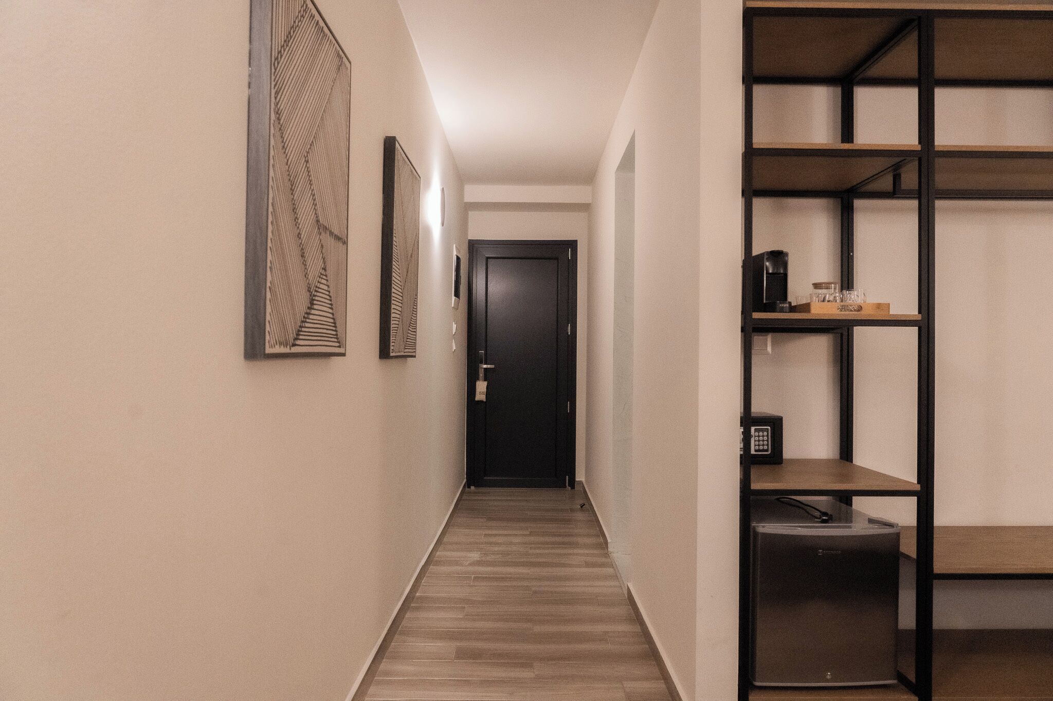A hallway with a wooden floor, a black door at the end, and a shelf unit with a coffee maker and a small refrigerator.