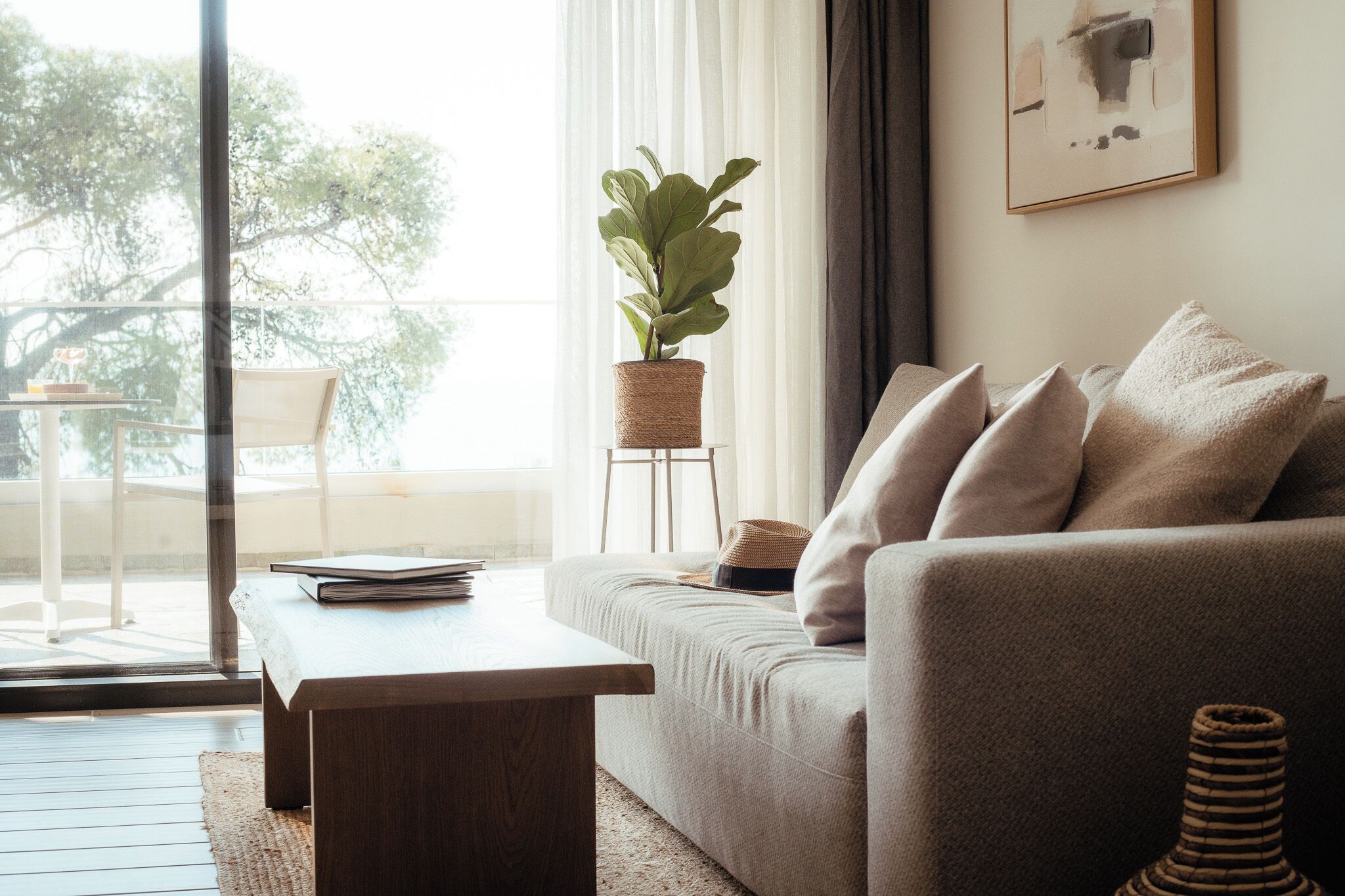A living room with a sofa, a coffee table, a potted plant, a framed picture on the wall, and a view of the outdoors.