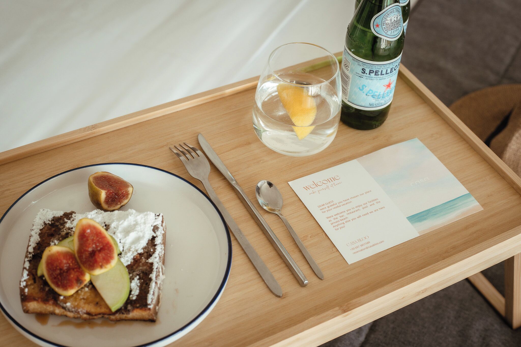 A wooden tray with a plate of food, a bottle of S. Pellegrino water, a glass with a lemon slice, and a welcome card.