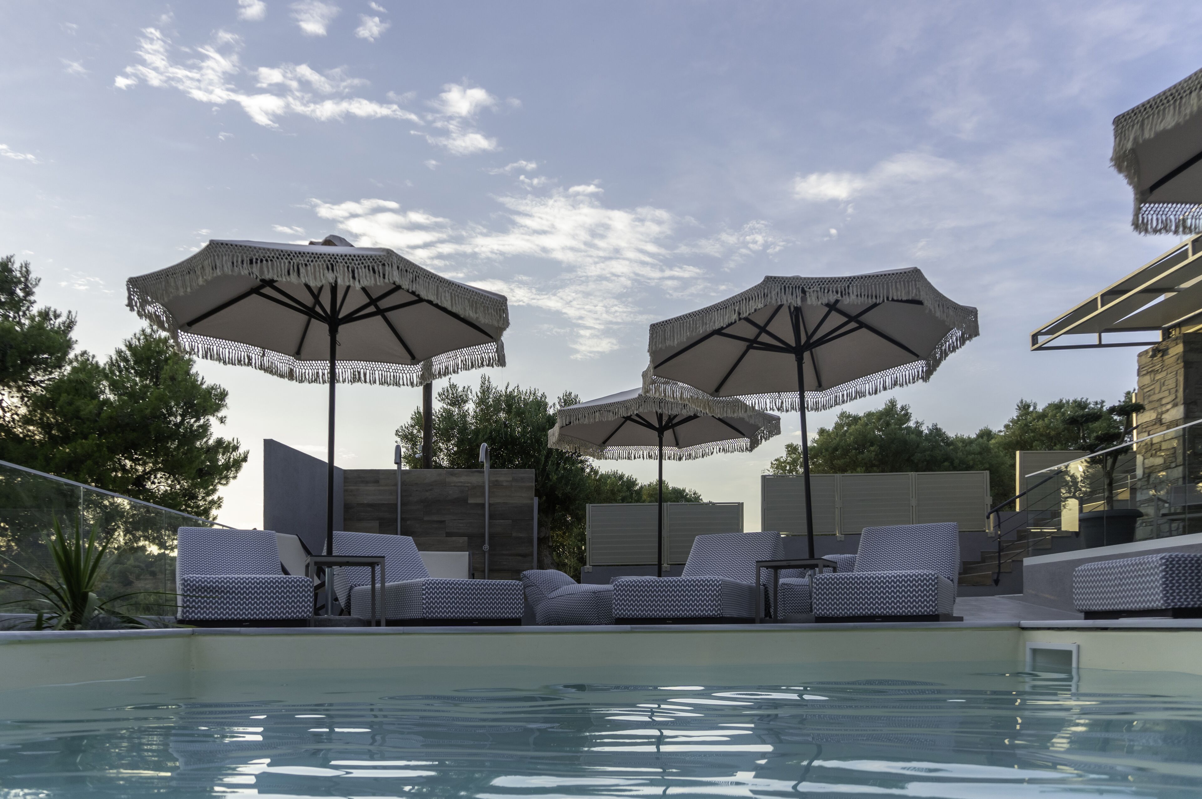 Zona junto a la piscina con sillones y sombrillas, rodeada por un muro de piedra y árboles.