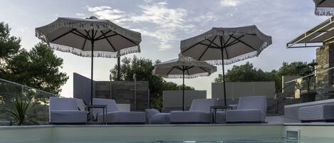 Zona junto a la piscina con sillones y sombrillas, rodeada por un muro de piedra y árboles.