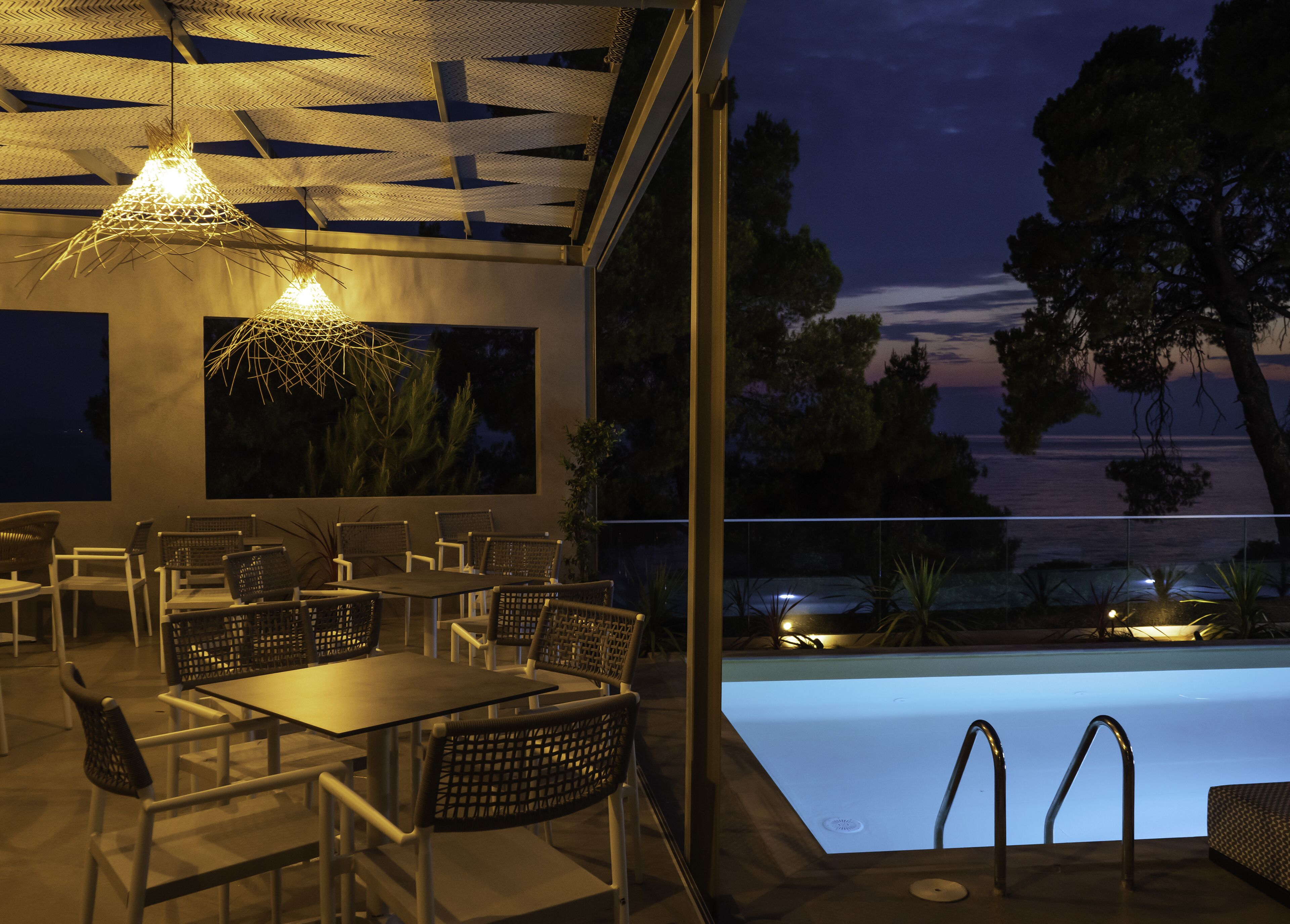 An outdoor dining area with tables and chairs, a pool, and a view of the sea at dusk.