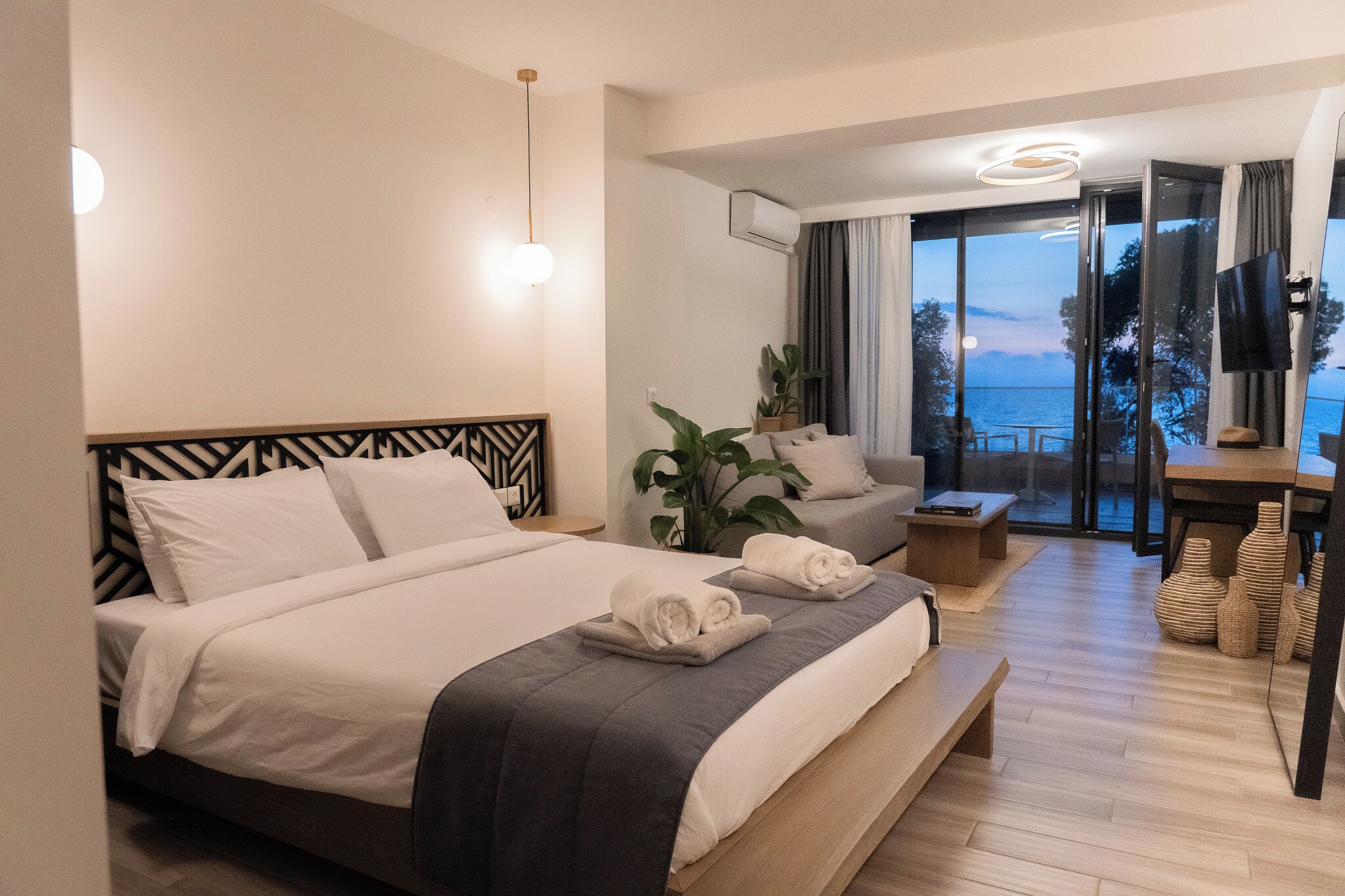 A modern hotel room with a large bed, a seating area with a view of the sea, and a balcony with a table and chairs.