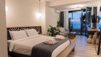 A modern hotel room with a large bed, a seating area with a view of the sea, and a balcony with a table and chairs.