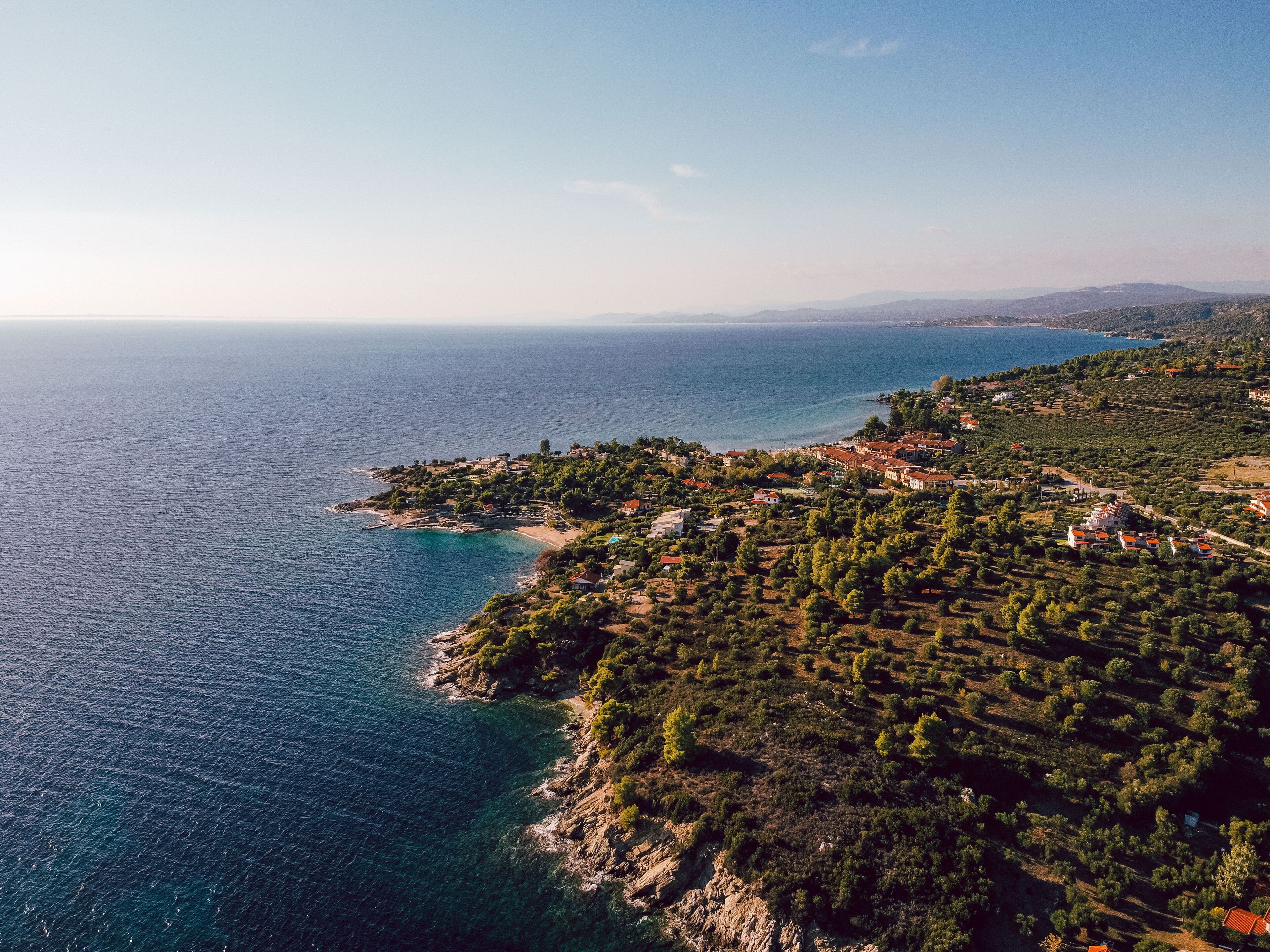 Vista aerea di una località costiera con case a schiera, un mare blu limpido e un paesaggio collinare.
