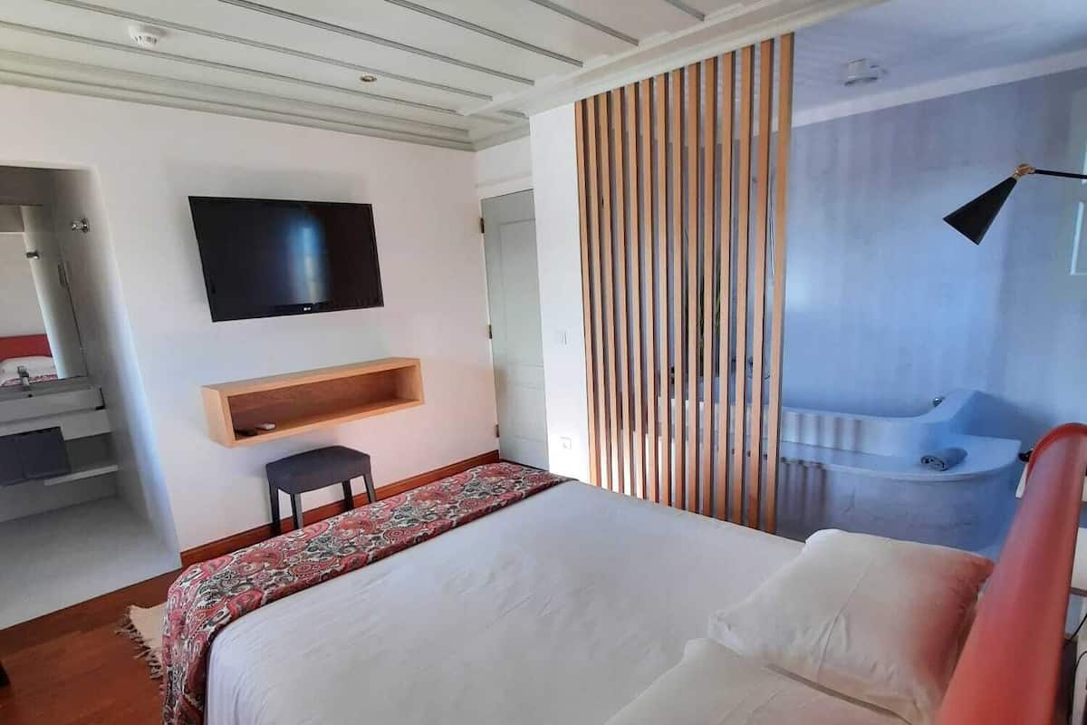 Une chambre moderne avec un grand lit, un téléviseur fixé au mur, une étagère en bois et une salle de bain avec une baignoire.