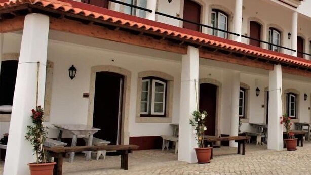 Un edificio con fachada blanca, detalles rojos y techo de tejas. Tiene varias ventanas y puertas, y el suelo está pavimentado con adoquines.