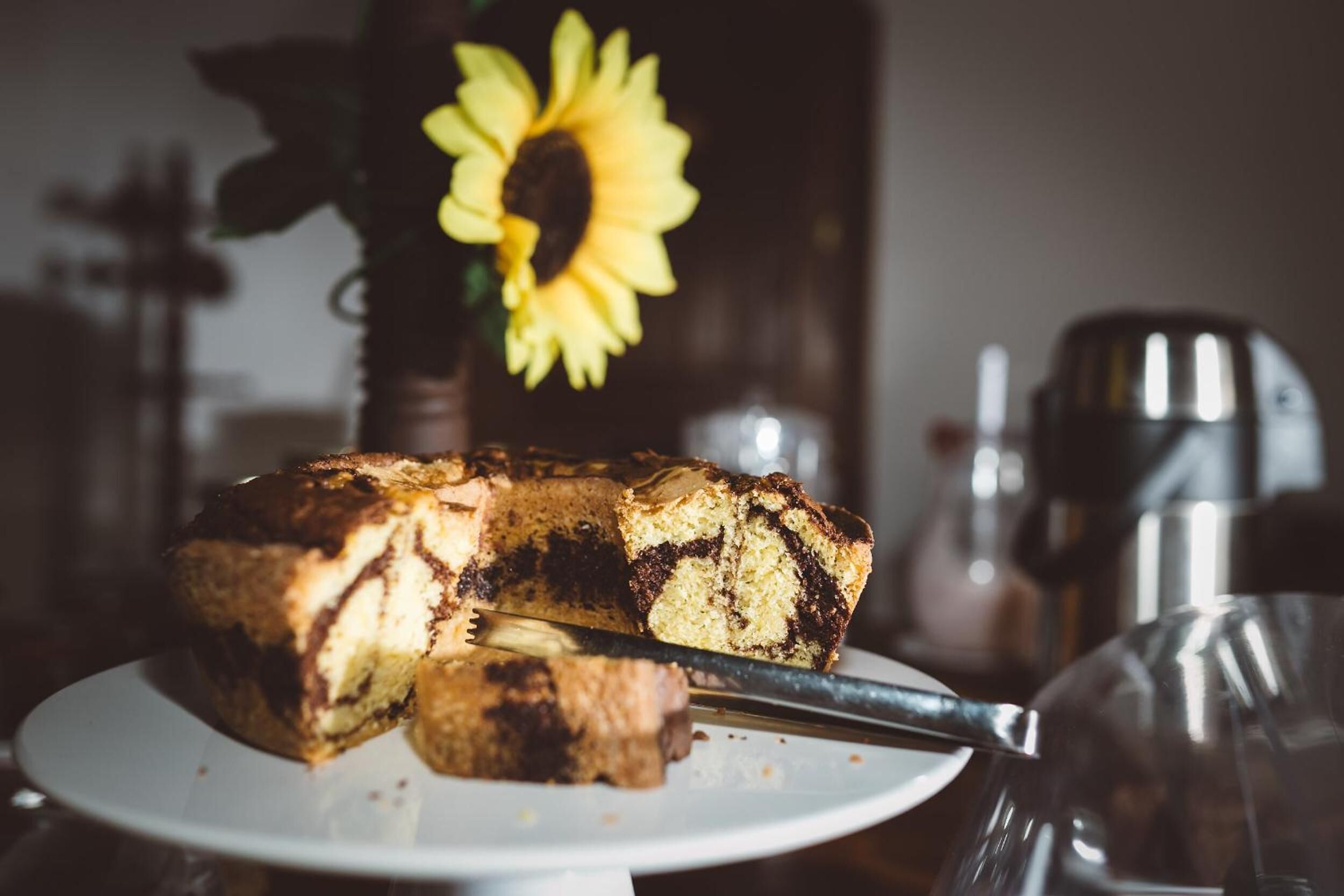 A slice of marble cake on a white plate with a knife, a sunflower in the background, and a coffee machine to the right.