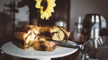 A slice of marble cake on a white plate with a knife, a sunflower in the background, and a coffee machine to the right.