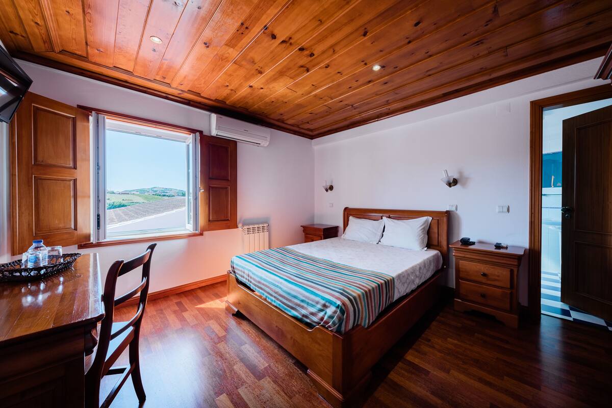 Une chambre avec un plafond en bois, une grande fenêtre, un lit recouvert d’une couverture à rayures, une table de chevet en bois et un bureau en bois avec une chaise.