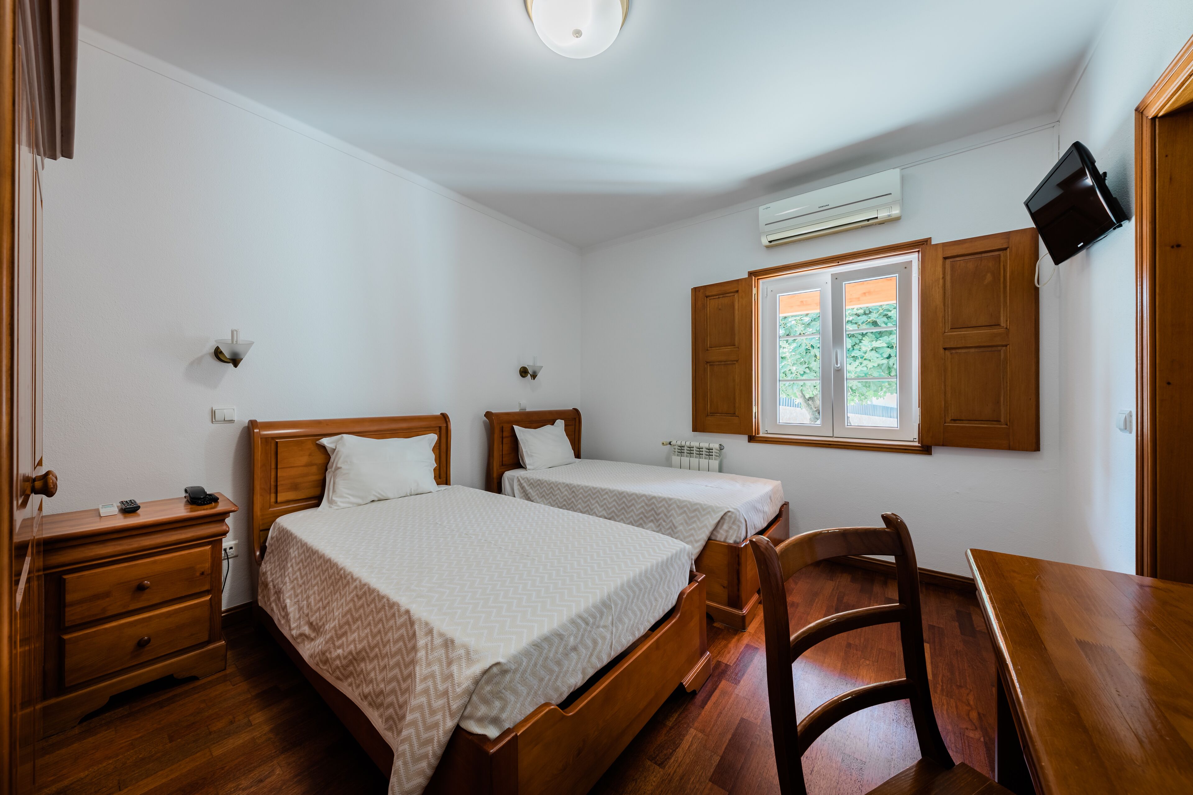 Ein Schlafzimmer mit zwei hölzernen Betten, einem hölzernen Schminktisch, einem Holzstuhl, einem Holztisch und einem Fenster mit Blick auf Grünflächen.