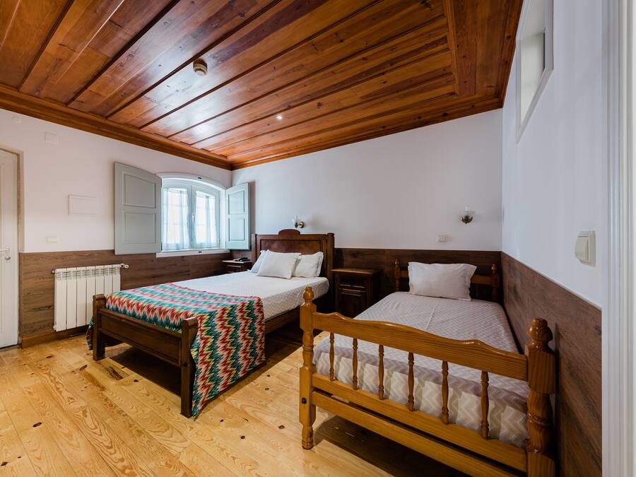 Un dormitorio con dos camas de madera, techo de madera, radiador blanco y ventana con vistas al exterior.