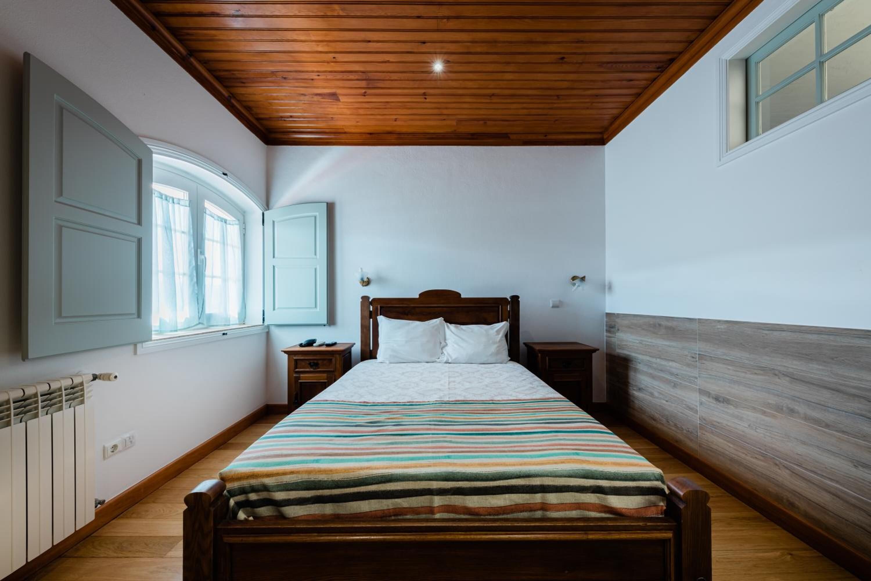 Un dormitorio con techo de madera, una cama con manta a rayas, dos mesitas de noche y una ventana con cortinas.