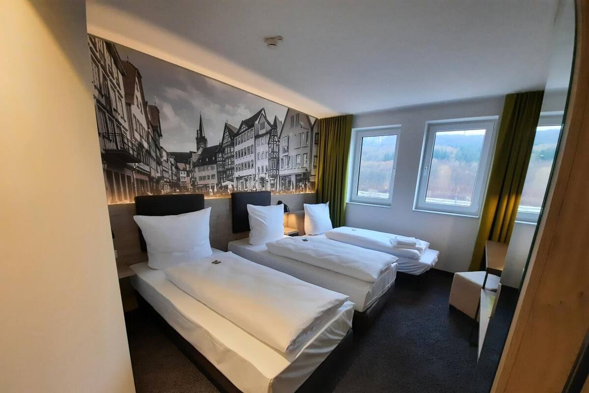 A hotel room with two beds, a mural of a European town on the wall, and a view of the city through the windows.
