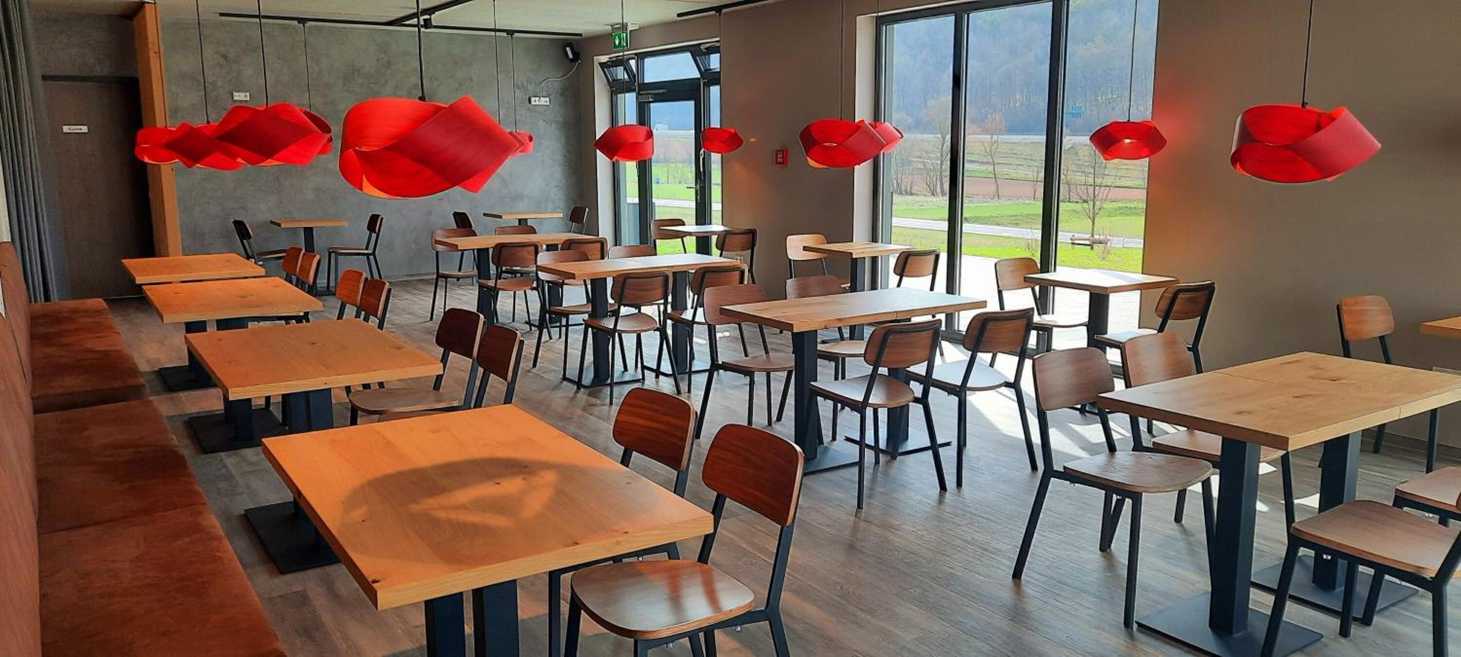 A modern dining area with wooden tables and chairs, large windows offering a view of the outdoors, and red pendant lights.