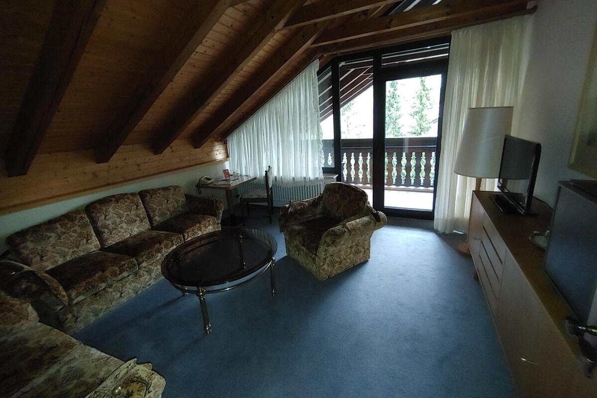 A room with a wooden ceiling, a sofa, a chair, a small table, a TV, and a window with curtains.