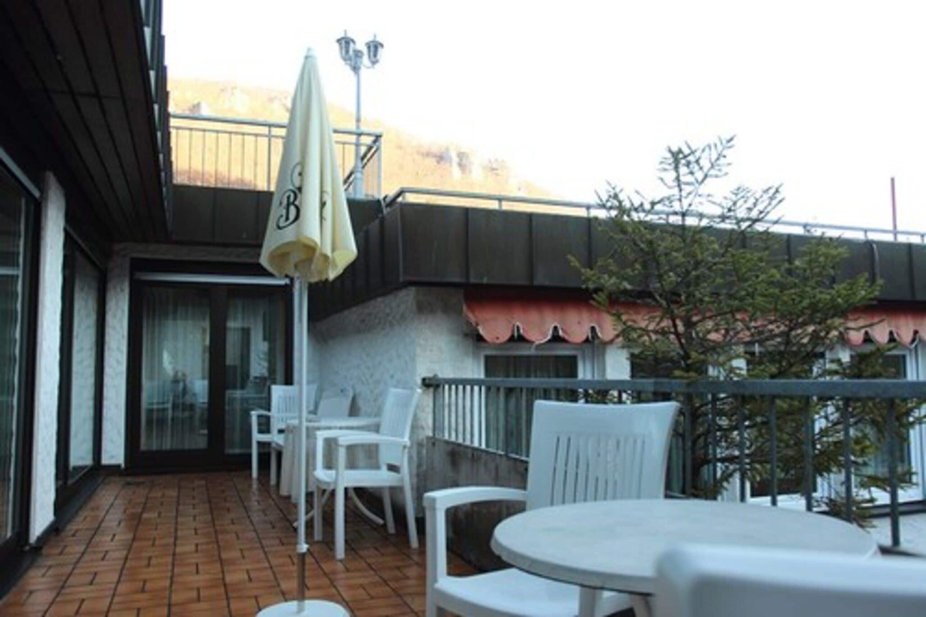 Un pati amb caires i taules de plàstic blanc, un petit parasol i la barana d'un balcó.