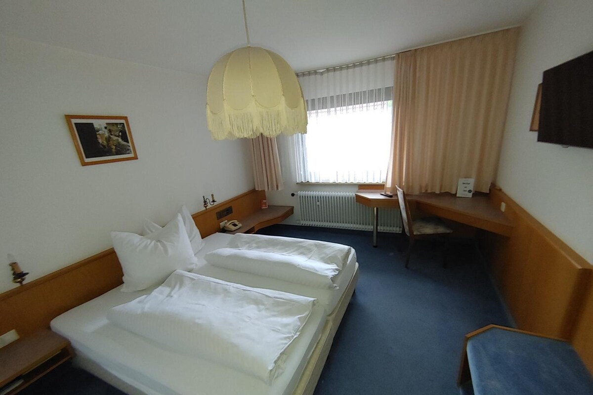 Habitación de hotel con cama doble, escritorio, silla, radiador y ventana con cortinas.