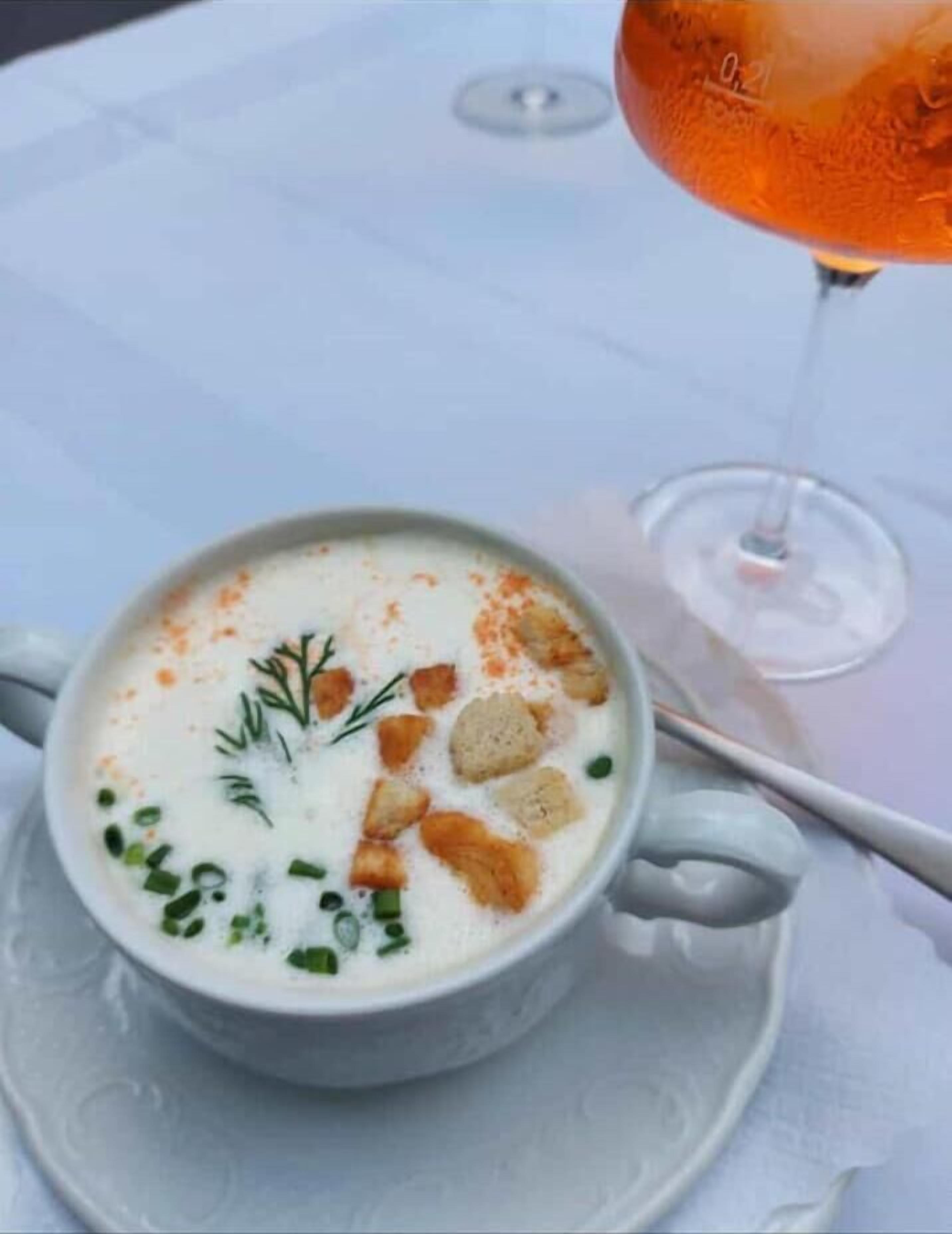 A white ceramic cup with a creamy soup garnished with herbs and croutons, accompanied by a glass of orange beverage on a white tablecloth.