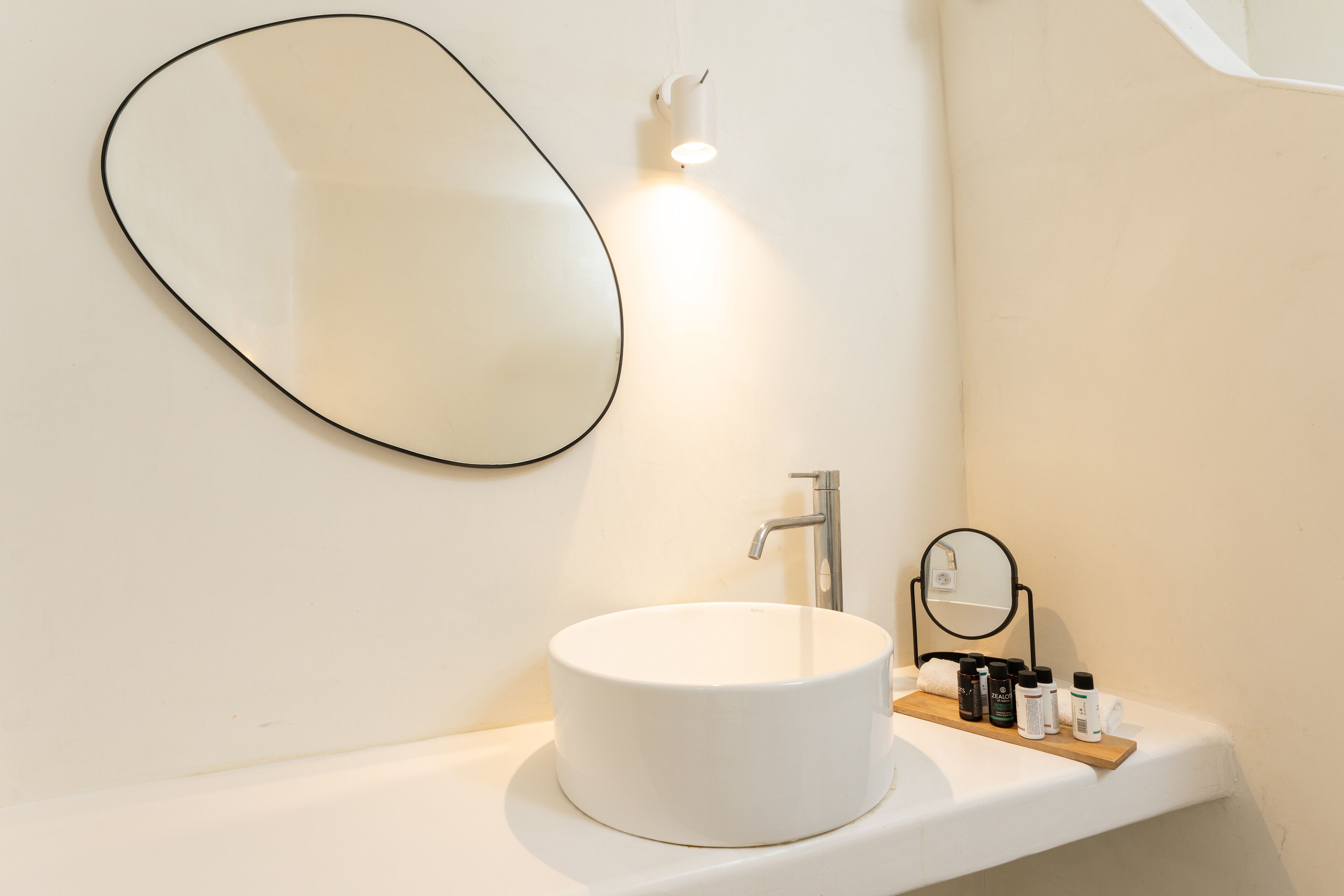 Un baño con un lavabo blanco redondo, un espejo ovalado grande y un espejo redondo pequeño sobre un estante de madera.